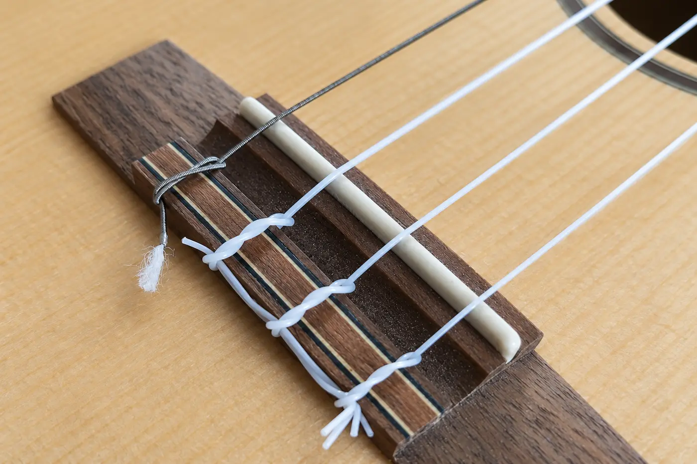 Umsponnene Low-G-Saite aus Metall am Steg einer Tenor-Ukulele neben drei weißen Kunststoffsaiten