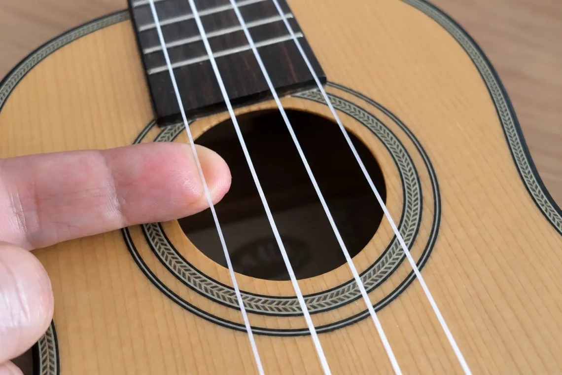 Ein Finger zupft eine Saite einer Konzert-Ukulele am Schallloch.